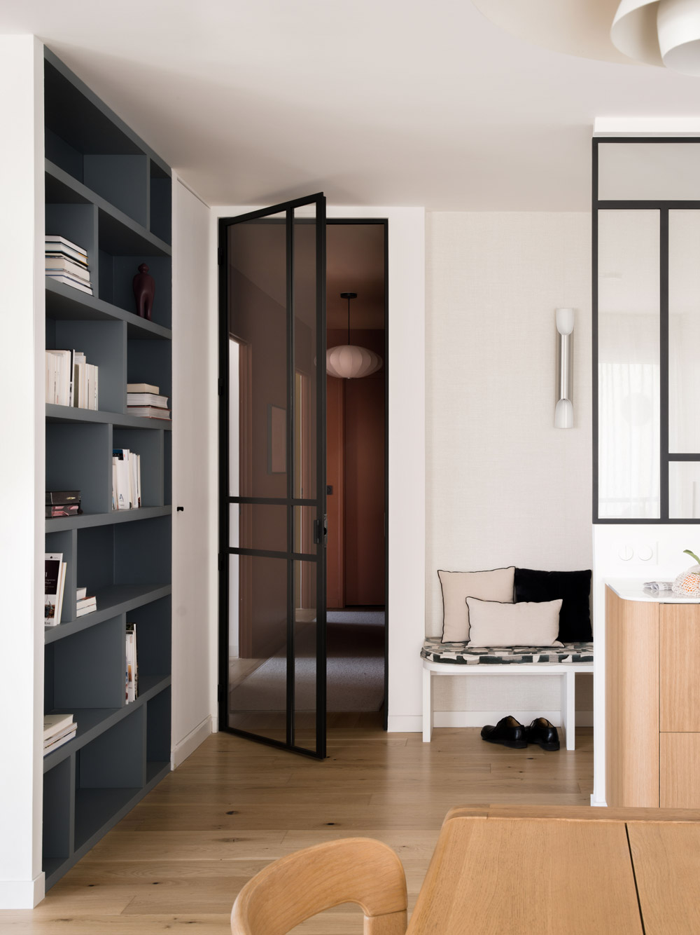 Passage entre bibliothèque, banquette et verrière noire dans un appartement rénové.