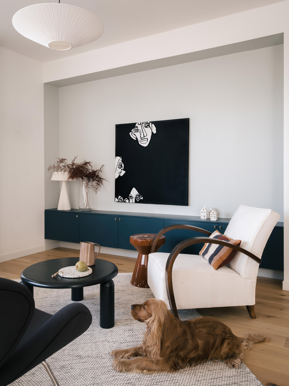 Salon avec meuble bas bleu, fauteuil blanc, tableau noir et chien sur tapis clair.