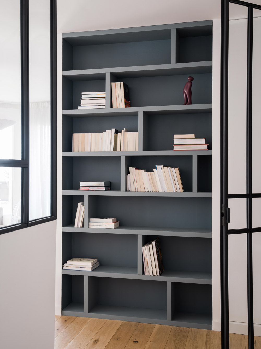 Bibliothèque sur mesure gris bleu encadrée par des verrières dans un appartement rénové.