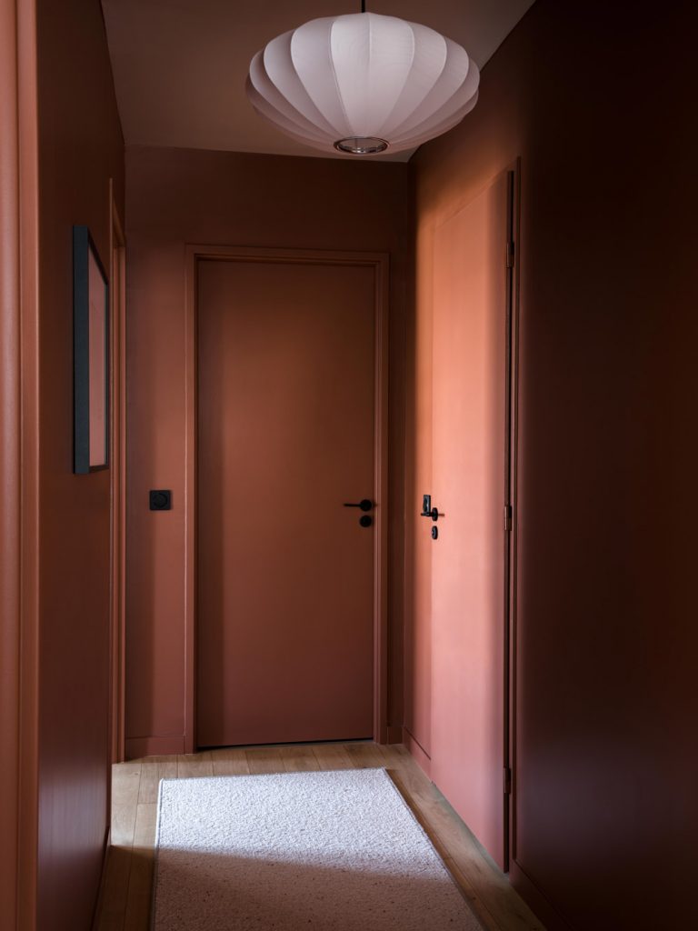 Couloir terracotta avec portes ton sur ton et suspension blanche dans l’appartement Simone Veil.