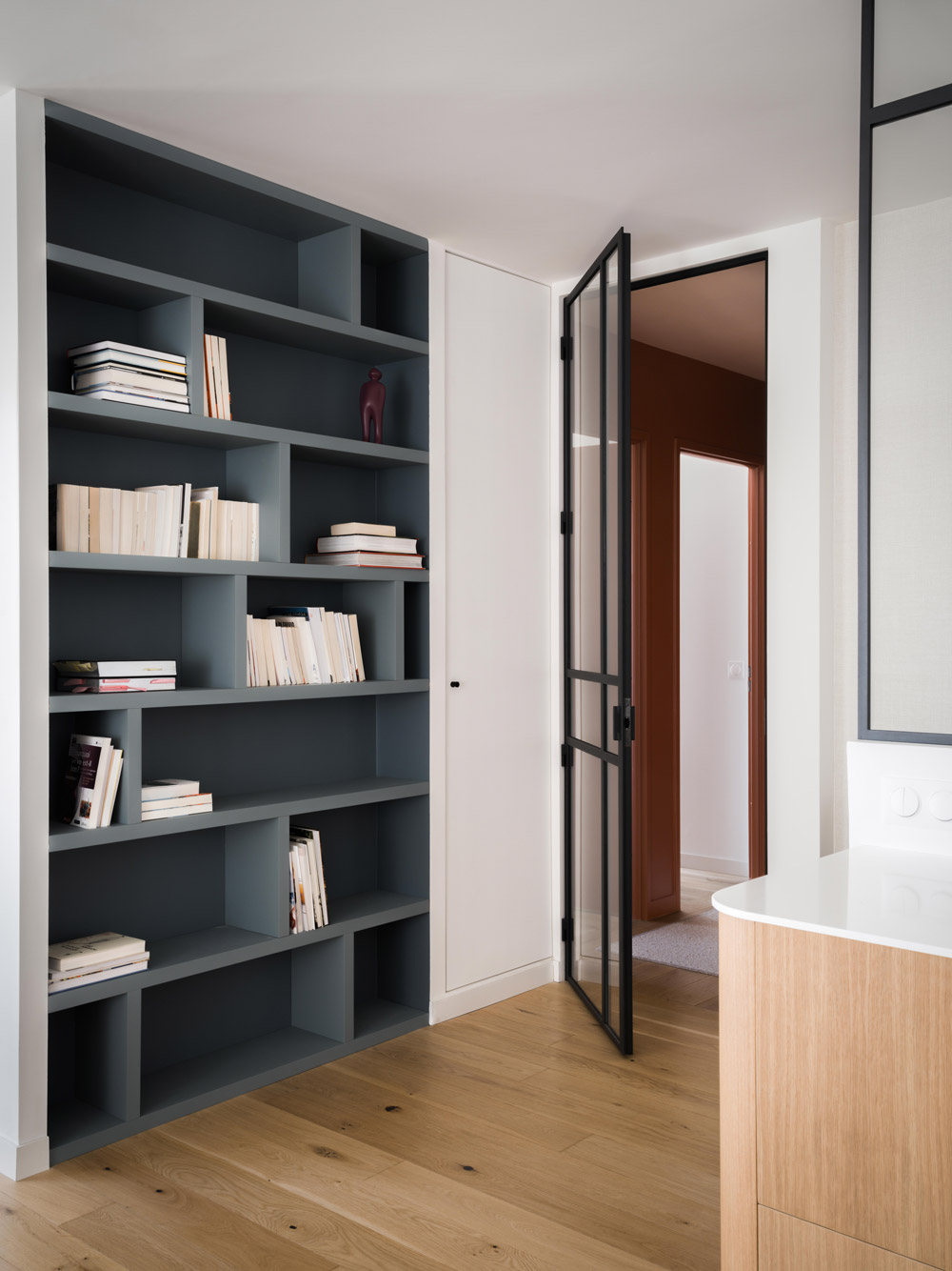 Porte vitrée noire ouverte sur un couloir terracotta près d’une bibliothèque sur mesure.