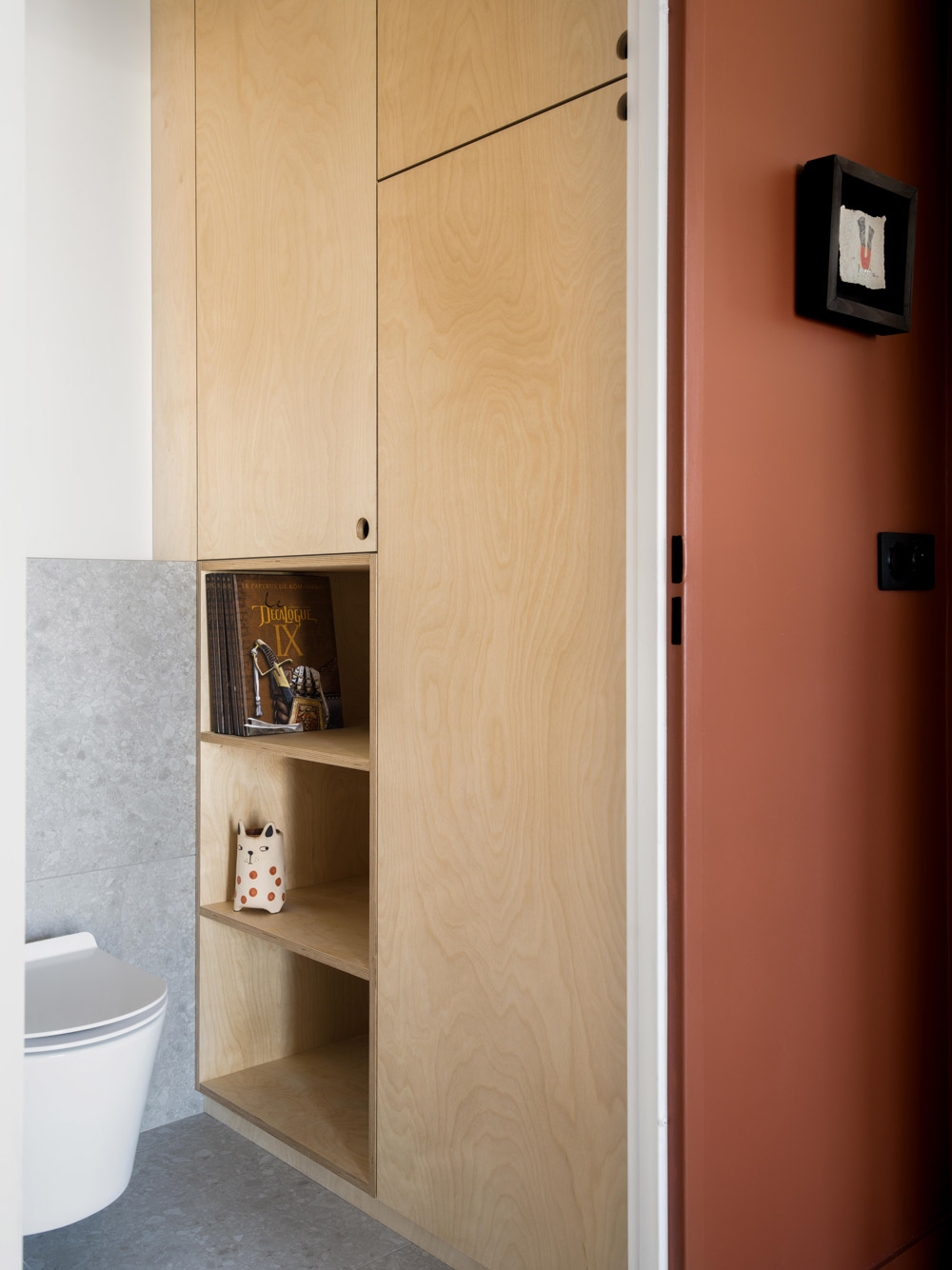 Rangements en bois clair dans une salle d’eau avec carrelage gris et couloir terracotta.